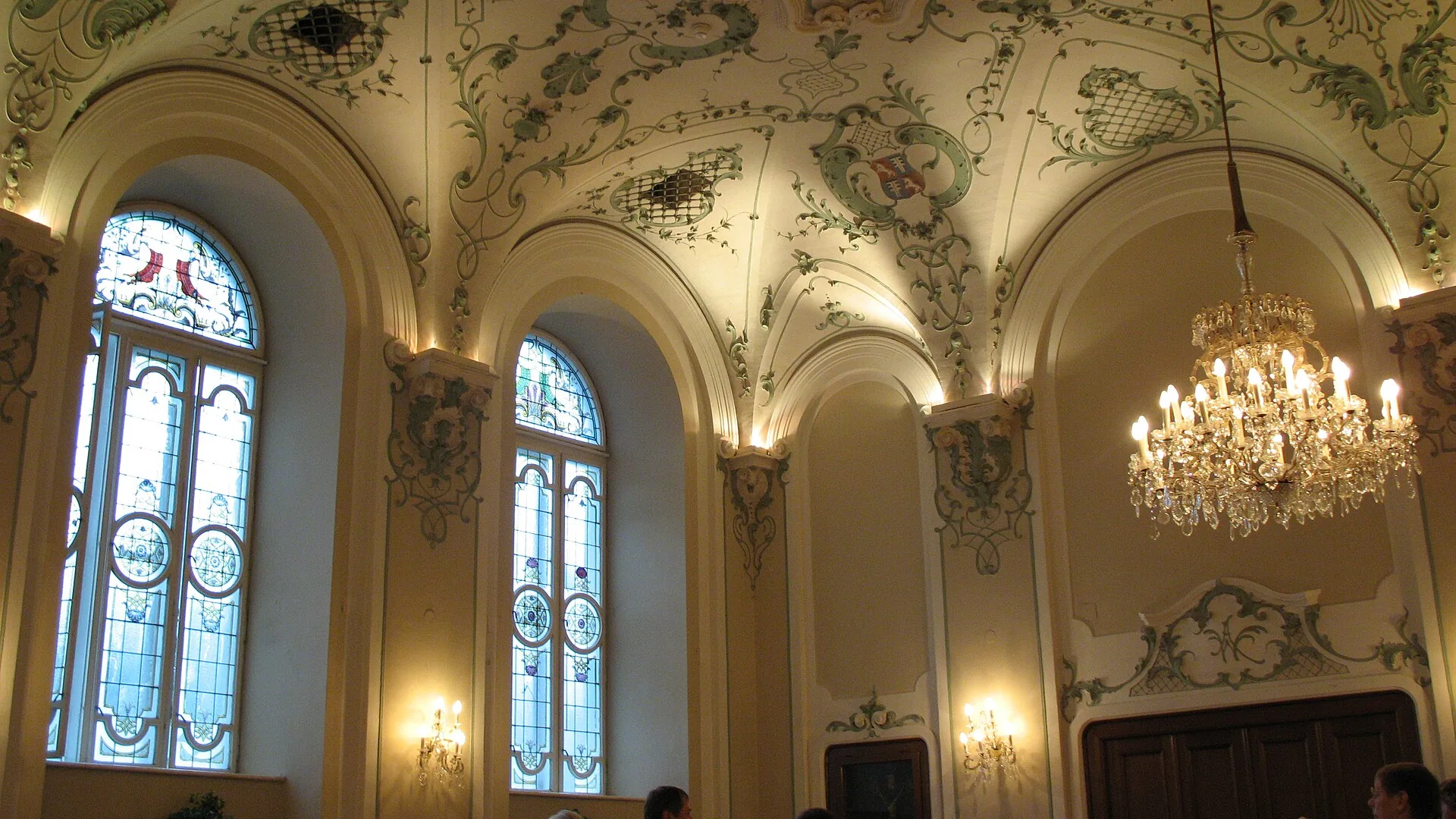A stuccoed hall of the St. Peter Stiftskeller in Salzburg, with arched windows, a crystal chandelier and a vaulted ceiling of green-and-white scrollwork.