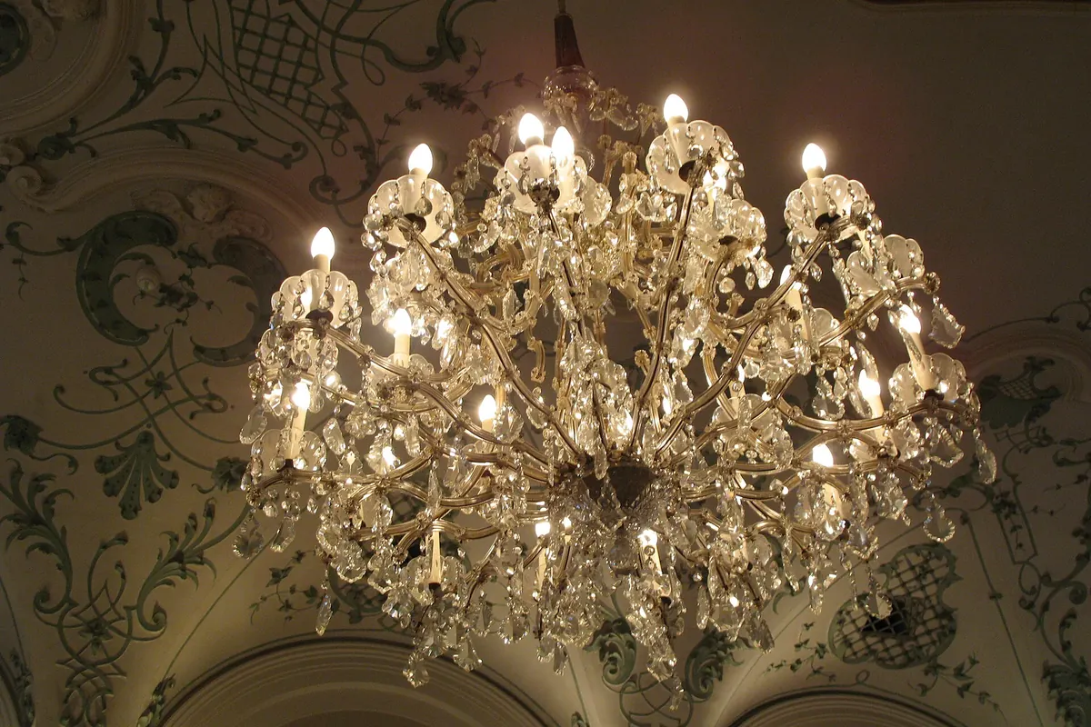 A crystal chandelier with candle-shaped bulbs hanging below a vaulted ceiling painted with green scrollwork.