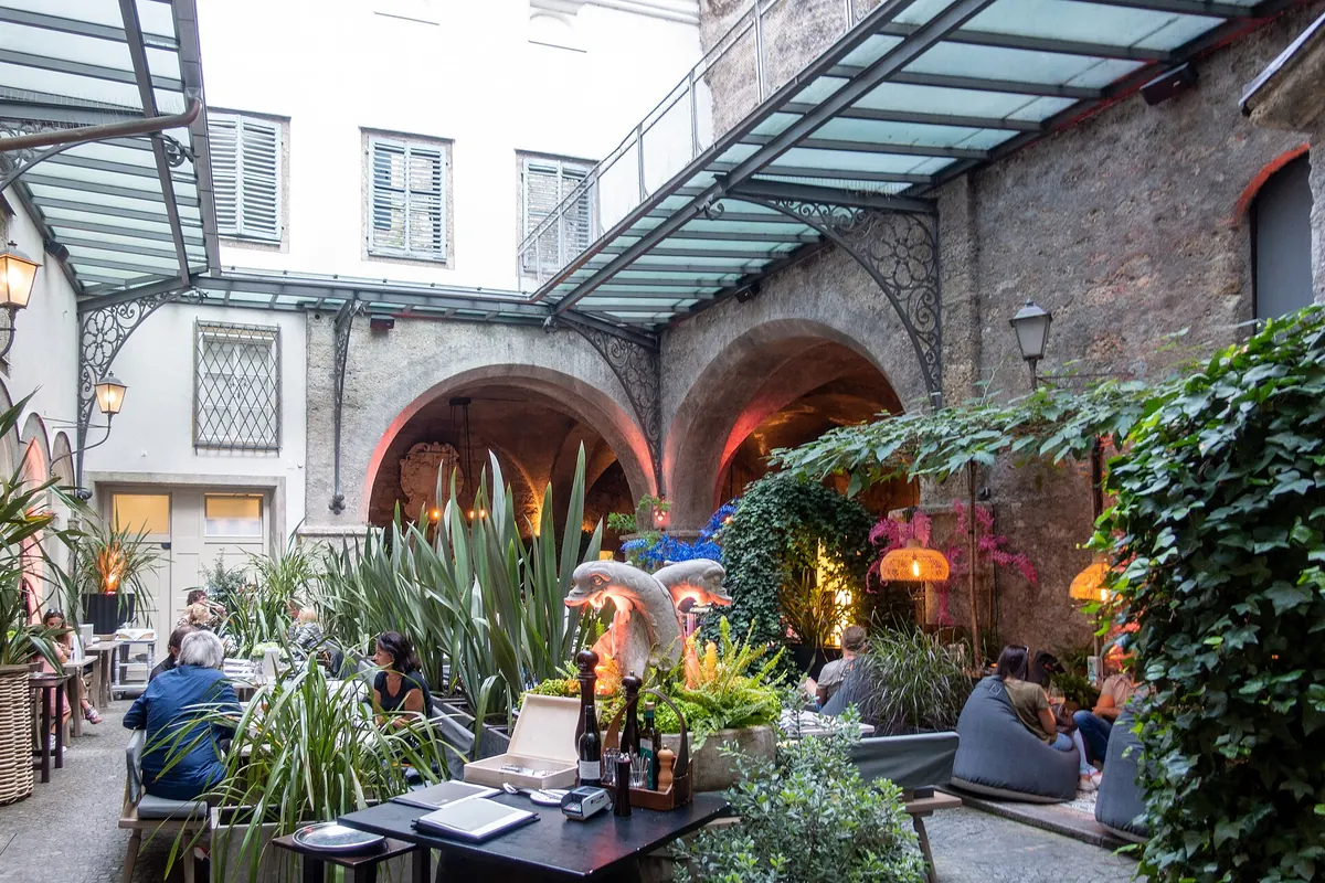 A courtyard of the St. Peter Stiftskulinarium with tables, plants and stone arches under a glass canopy.